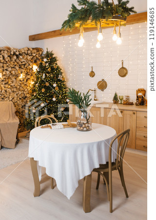 Round table with a white tablecloth and a vase with fir branches stand in the kitchen near the Christmas tree Round table with a white tablecloth and a vase with fir branches stand in the kitchen near the Christmas tree 119146685