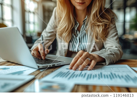 Office worker woman is sitting at a desk with a laptop and a stack of papers and writing on the papers with a pen, working on a project or a report 119146813