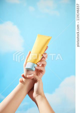 Under a blue sky with a few clouds and strong light, a hand is holding the wrist of another hand holding an unbranded yellow tube of cream with a blue cap, ideal for illustrating skincare content. Under a blue sky with a few clouds and strong light, a hand is holding the wrist of another hand holding an unbranded yellow tube of cream with a blue cap, ideal for illustrating skincare content. 119146837