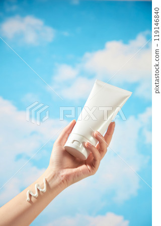 In the photo, used to advertise a skincare product, the background is a beautiful blue sky with moving clouds. A female model is holding an unbranded white tube with a smear of cream on her wrist. 119146840