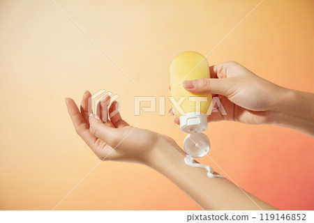 The minimalist composition image features a model hand squeezing a yellow tube with a white cap off a cream piece, a bright orange background resembling the sun, suitable for sunscreen commercials. 119146852