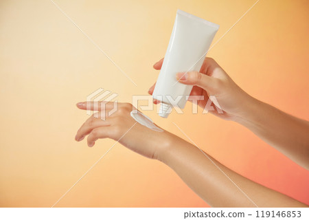 The female model gently squeezes a sample of cream onto her other hand from an unlabeled white tube under a bright orange background, suitable for a sunscreen advertisement. 119146853