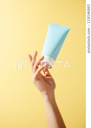 An up-close picture shows a blue squeeze bottle with a white top suspended in midair. A lady reaches up to take the bottle. The backdrop has a vivid orange color. 119146865