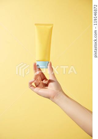 A model hand gently grips an unlabeled bright yellow tube of cream with the blue cap against a sunny yellow background, creating a pop of color, perfect for skincare advertisements. 119146872