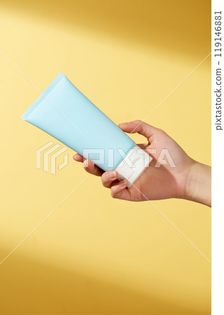 A female hand holds an unlabeled blue tube of lotion against a bright yellow background, illuminated by sunshine. This simple yet elegant composition showcases the product. 119146881