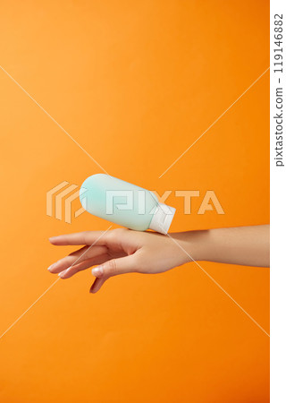 Minimalist layout image for sunscreen product advertisement on a bright orange background like the sun features an unlabeled blue bottle gently floating down onto the back of an outstretched hand. 119146882