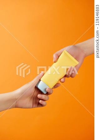 Two hands are holding an unlabeled yellow tube of cream from both ends of the tube, creating an attractive feeling from the tube, perfect for advertising sunscreen against a bright orange background. 119146883