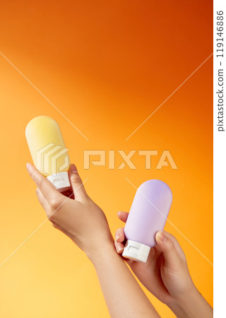 A hand is gripping the cap of the same mockup cream tube without the purple and yellow label in this minimalist picture layout for an advertisement for a skincare product on an orange backdrop. A hand is gripping the cap of the same mockup cream tube without the purple and yellow label in this minimalist picture layout for an advertisement for a skincare product on an orange backdrop. 119146886