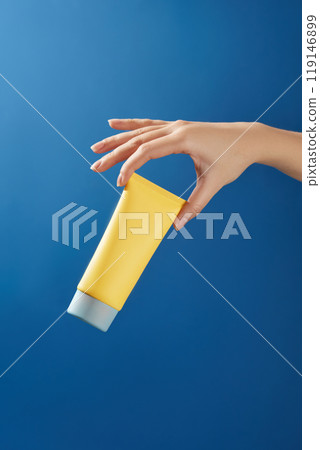 A model hand gently lifts the bottom of an unbranded yellow tube of cream and shows it in a dark blue background resembling a sunny sky, creating the premise for a template of sunscreen advertisement. 119146899