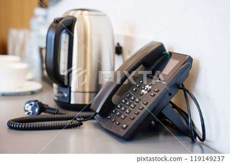 A corded black telephone in the hotel room on a table with a kettle and cups A corded black telephone in the hotel room on a table with a kettle and cups 119149237