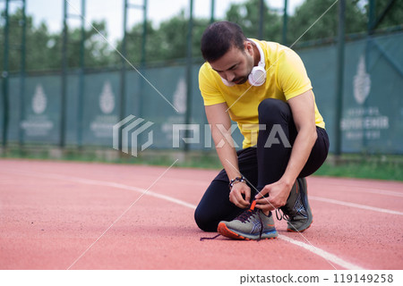 Athlete tying sneaker laces while squatting on rubberized running track during training break at urban outdoor arena 119149258