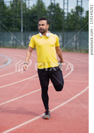 Vibrant athlete in wireless headphones warms up leg muscles on rubberized running track at city park's sports ground. 119149261