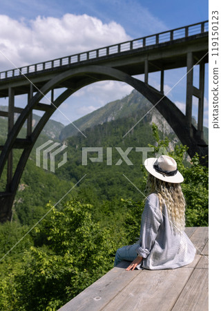A girl in a hat stands against the backdrop of a beautiful arched bridge over the Tara River, a popular tourist attraction in Montenegro. 119150123