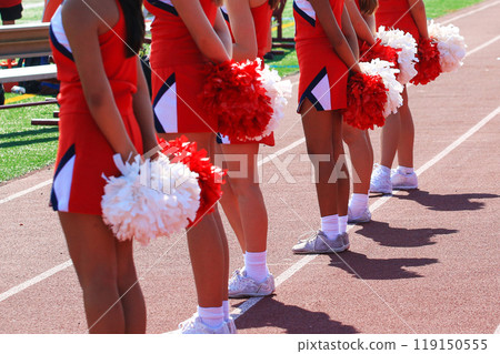 Cheerleaders holding their pom poms behind their backs on the sidelines of a football game 119150555