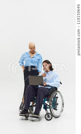Young bald woman in blue shirt standing next to man in wheelchair working on laptop, showcasing inclusivity and collaboration in workplace. 119150649