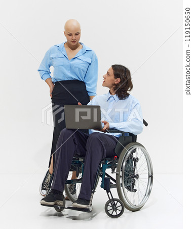 Young bald woman in blue shirt standing next to man in wheelchair working on laptop, showcasing inclusivity and collaboration in workplace. 119150650
