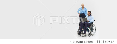 Young bald woman in blue shirt standing next to man in wheelchair working on laptop, showcasing inclusivity and collaboration in workplace. 119150652