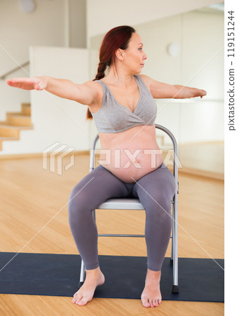 Pregnant woman is engaged in yoga. Exercise on a chair with arms spread apart Pregnant woman is engaged in yoga. Exercise on a chair with arms spread apart 119151244