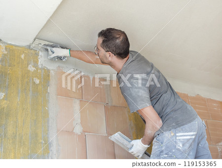 Craftsman in dirty gloves and gray shirt spreading plaster with a metal float over a ceiling Craftsman in dirty gloves and gray shirt spreading plaster with a metal float over a ceiling 119153365