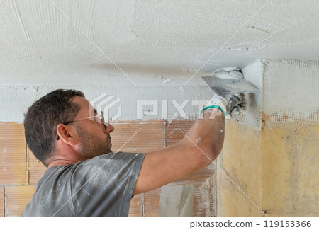 Craftsman in dirty gloves and gray shirt spreading plaster with a metal float over a ceiling Craftsman in dirty gloves and gray shirt spreading plaster with a metal float over a ceiling 119153366