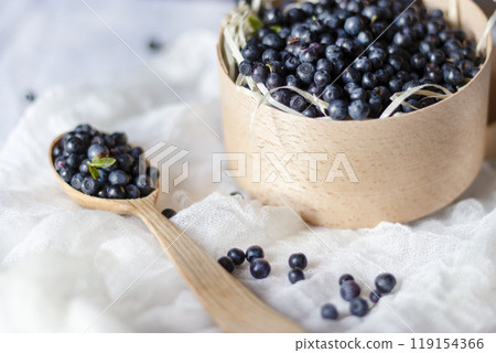 Blueberries in a wooden bowl, next to a wooden spoon with berries, lie on a white canvas, still life with wooden box with bilberries. Blueberries in a wooden bowl, next to a wooden spoon with berries, lie on a white canvas, still life with wooden box with bilberries. 119154366