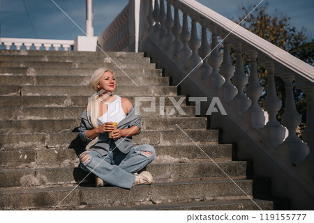 A woman sits on a set of stairs, holding a cup of coffee. She is enjoying her coffee and taking in the view from the top of the stairs. 119155777