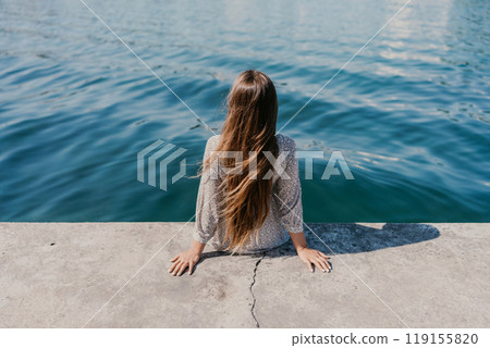 A woman is sitting on a ledge by a body of water, wearing a dress with a leopard print pattern. The scene is peaceful and serene, with the woman enjoying the view of the water and the surrounding area 119155820