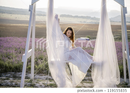 A woman is sitting on a bench under a white canopy. She is wearing a white dress and has long hair. The scene is set in a field with purple flowers. A woman is sitting on a bench under a white canopy. She is wearing a white dress and has long hair. The scene is set in a field with purple flowers. 119155835