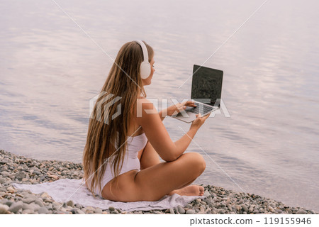 A woman is sitting on a beach with a laptop in her lap. She is wearing headphones and she is listening to music. A woman is sitting on a beach with a laptop in her lap. She is wearing headphones and she is listening to music. 119155946