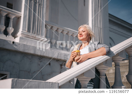 A woman is standing on a white railing with a cup of coffee in her hand. She is wearing a gray jacket and blue jeans. Concept of relaxation and leisure. 119156077