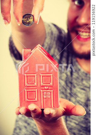 Smiling man putting coin into house piggybank Smiling man putting coin into house piggybank 119156922