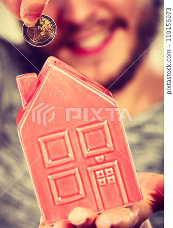 Smiling man putting coin into house piggybank 119156973