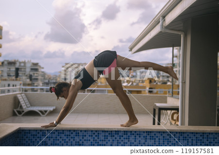 A Young Woman Engaged in Practicing a Yoga Pose Beside the Pool Area Under the Sun A Young Woman Engaged in Practicing a Yoga Pose Beside the Pool Area Under the Sun 119157581