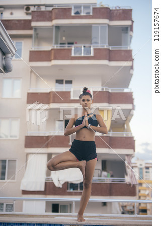 A woman engaging in the practice of yoga by a pool, surrounded by a vibrant urban setting A woman engaging in the practice of yoga by a pool, surrounded by a vibrant urban setting 119157674