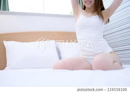 A young, slender woman in underwear stretches in her bedroom after waking up, basking in the morning sun 119158100