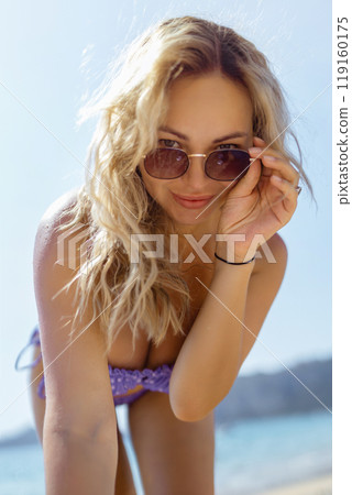 A beautiful girl in sunglasses on the beach looks at the camera, leaning towards it. Sexy blonde in a swimsuit on the seashore A beautiful girl in sunglasses on the beach looks at the camera, leaning towards it. Sexy blonde in a swimsuit on the seashore 119160175
