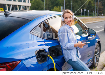 Young woman wearing light casual clothes relaxes with headphones, checking her phone as her electric car charges. Young woman wearing light casual clothes relaxes with headphones, checking her phone as her electric car charges. 119160338