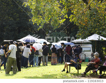 廚房車在園區的活動場地排隊。人們撐著遮陽傘，整齊地排列著。有人在柿子下乘涼休息。 119163013