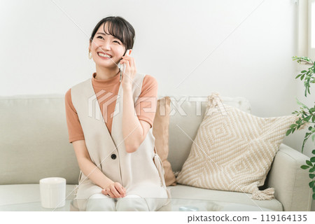 微笑的亞洲女人坐在沙發上,用智能手機給朋友打電話 微笑的亞洲女人坐在沙發上,用智能手機給朋友打電話 119164135