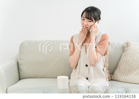 An Asian woman sitting on a sofa who is surprised and surprised while calling, calling, and talking on a smartphone 119164138