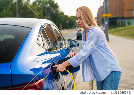 Blonde woman in casual clothes plugs her electric car into a charger at a public charging station Blonde woman in casual clothes plugs her electric car into a charger at a public charging station 119166978