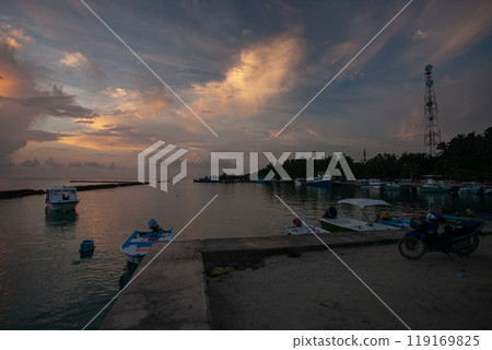 Tranquil sunset by the harbor in the Maldives, as boats rest gently in calm waters and clouds paint the sky Tranquil sunset by the harbor in the Maldives, as boats rest gently in calm waters and clouds paint the sky 119169825