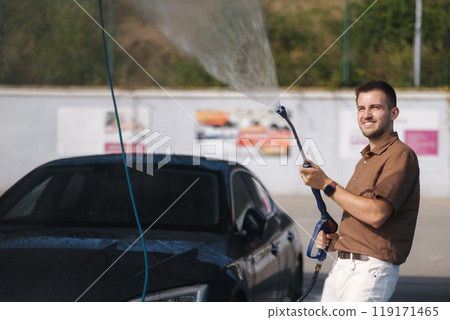 Happy young man at a car wash uses a pressure washer and sprays water uphill. Man is having fun Happy young man at a car wash uses a pressure washer and sprays water uphill. Man is having fun 119171465