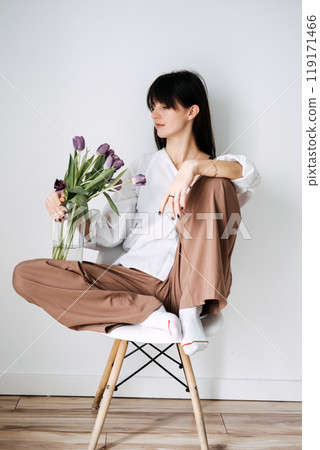 Young woman holding purple tulips while sitting cross-legged on a chair, minimalist indoor setting, floral and lifestyle concept 119171466