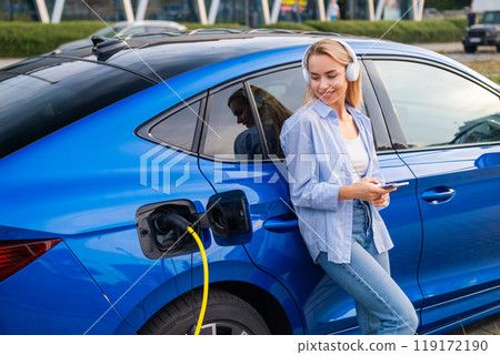 Blonde woman checks electric car charge level using phone while listening to music through headphones.  119172190
