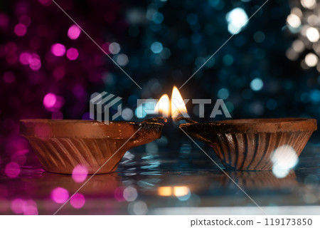 Two illuminated traditional clay diyas with glowing flames on a vibrant bokeh background of colorful pink and blue colors. 119173850
