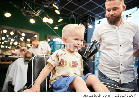 Cheerful child in barbershop chair at the end of haircut, barber using hairdryer to remove loose hair clippings. Enjoyment or satisfaction with the haircut experience. 119175167