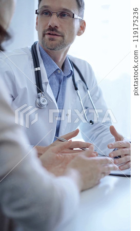 Doctor and a patient. The physician, wearing a white medical coat over a blue shirt, gesturing with his hands during a consultation in the clinic. Medicine concept 119175236