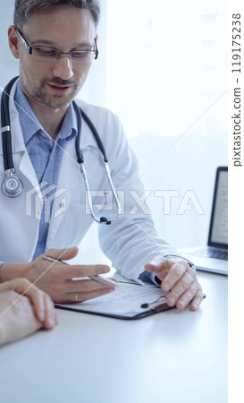 Doctor and a patient. The physician, wearing a white medical coat over a blue shirt, gesturing with his hands during a consultation in the clinic. Medicine concept 119175238