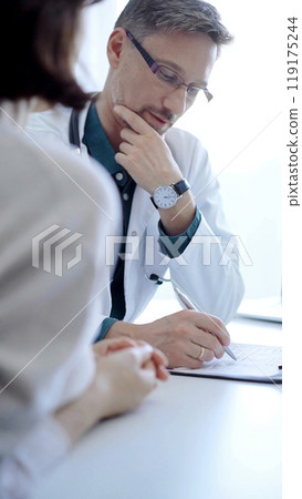 Doctor and a patient. The physician, wearing a white medical coat over a green shirt, gesturing with his hands during a consultation in the clinic. Medicine concept Doctor and a patient. The physician, wearing a white medical coat over a green shirt, gesturing with his hands during a consultation in the clinic. Medicine concept 119175244